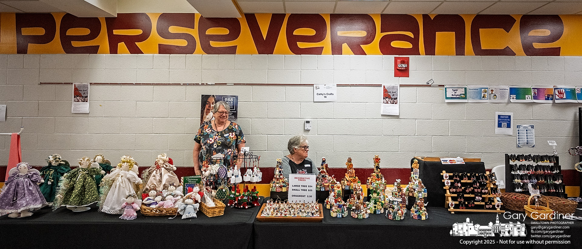 Two women sell handmade dolls and decorative trees in the main hallway by the student lockers at Westerville North High School during the Mark Twain Craft Bazaar. Above them, a banner reading "Perseverance," one of the school’s core values, underscores the spirit behind their craft.