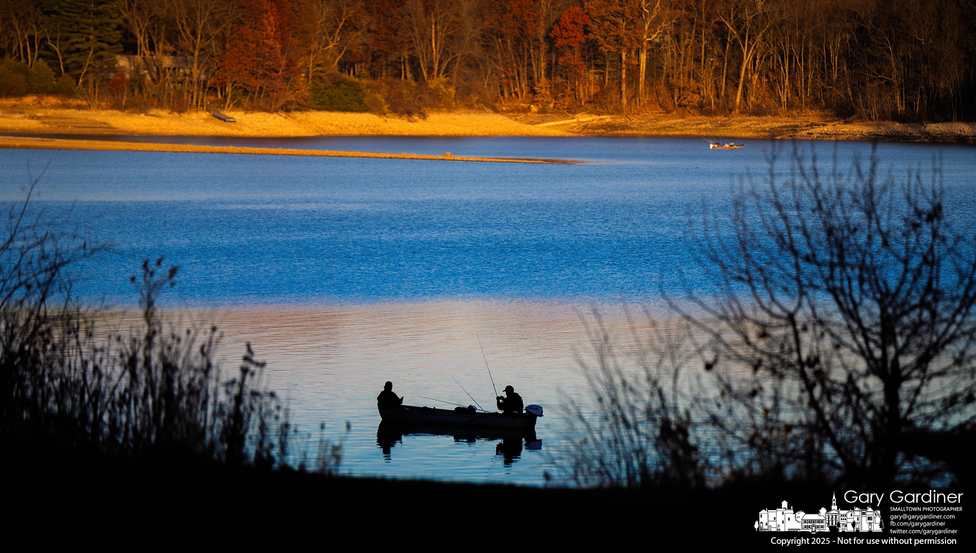 Two fishermen move their boat through the shadowed shallows of Hoover Reservoir as the setting sun casts a warm glow on the trees across the water. My Final Photo for November 23, 2025.
