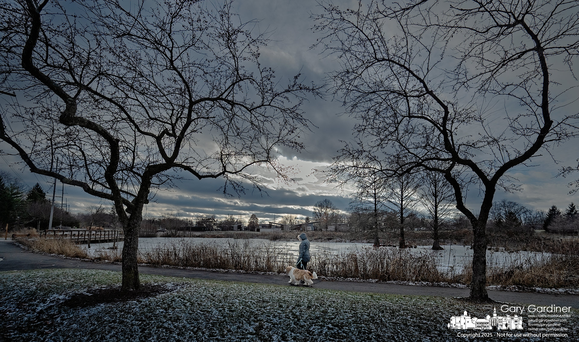 A woman walks her dog along the path past the pool at the Highlands wetlands as the sun begins to break through the clouds at sunset. My final Photo for December 30, 2025.