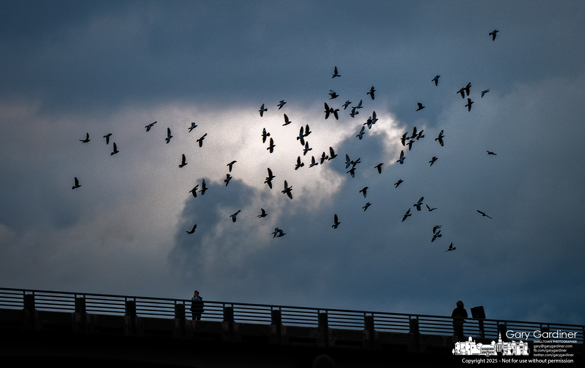 Onlookers pause along the railing as a flock of birds cuts through a bright opening in the clouds above Hoover Dam