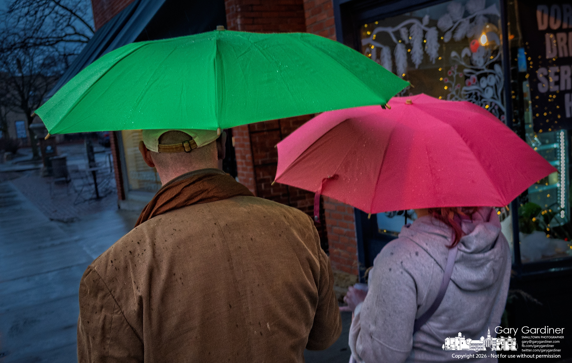 A couple walks side by side under green and pink umbrellas on a rainy afternoon in Uptown Westerville, with a steady, light rain and overcast skies giving the streets a quiet, reflective mood. My Final Photo for January 10, 2026.