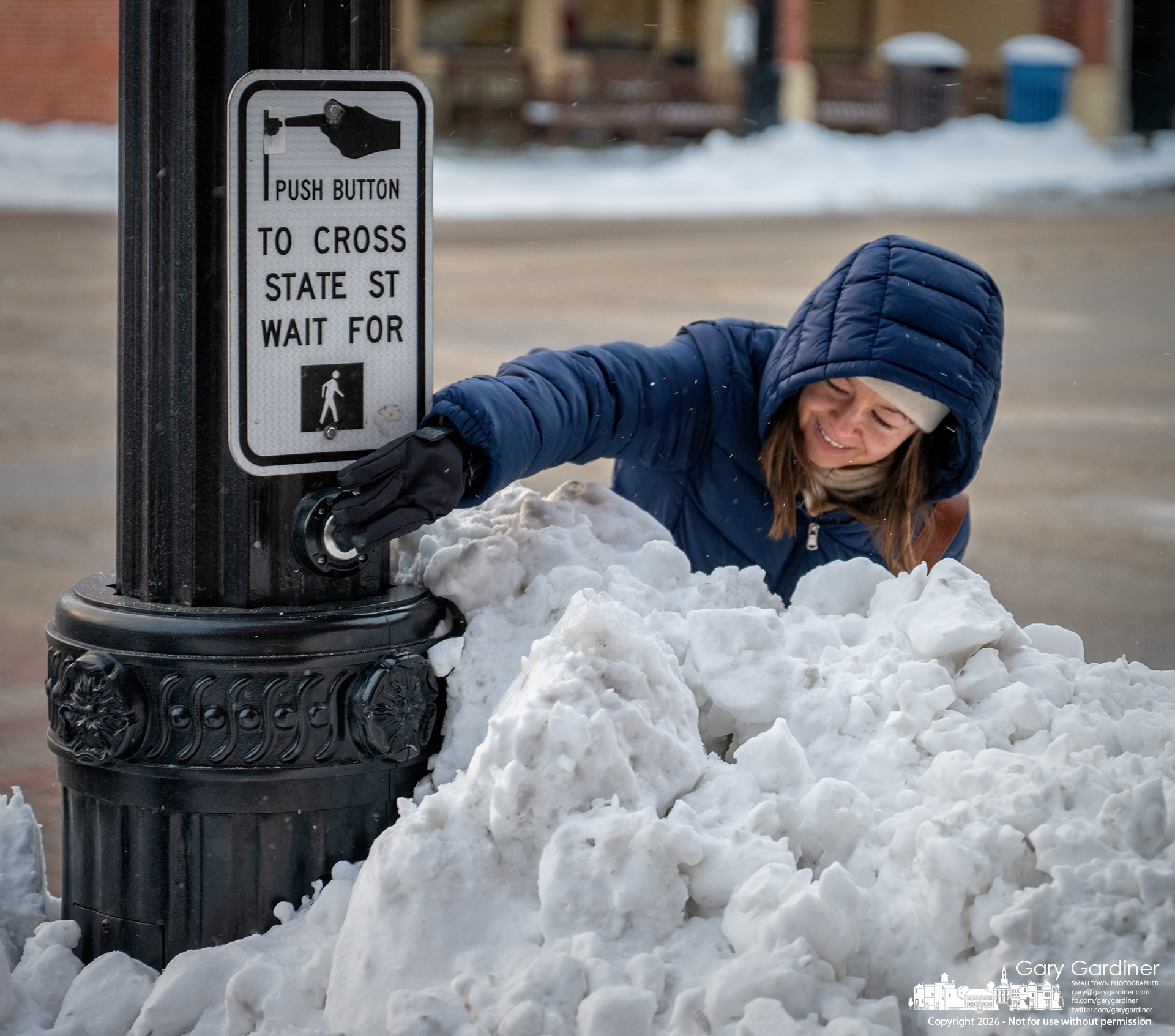 A woman stretches across a chest-high pile of plowed snow to activate the pedestrian crossing light at State and college Tuesday afternoon. (My Final Photo for January 27, 2026)