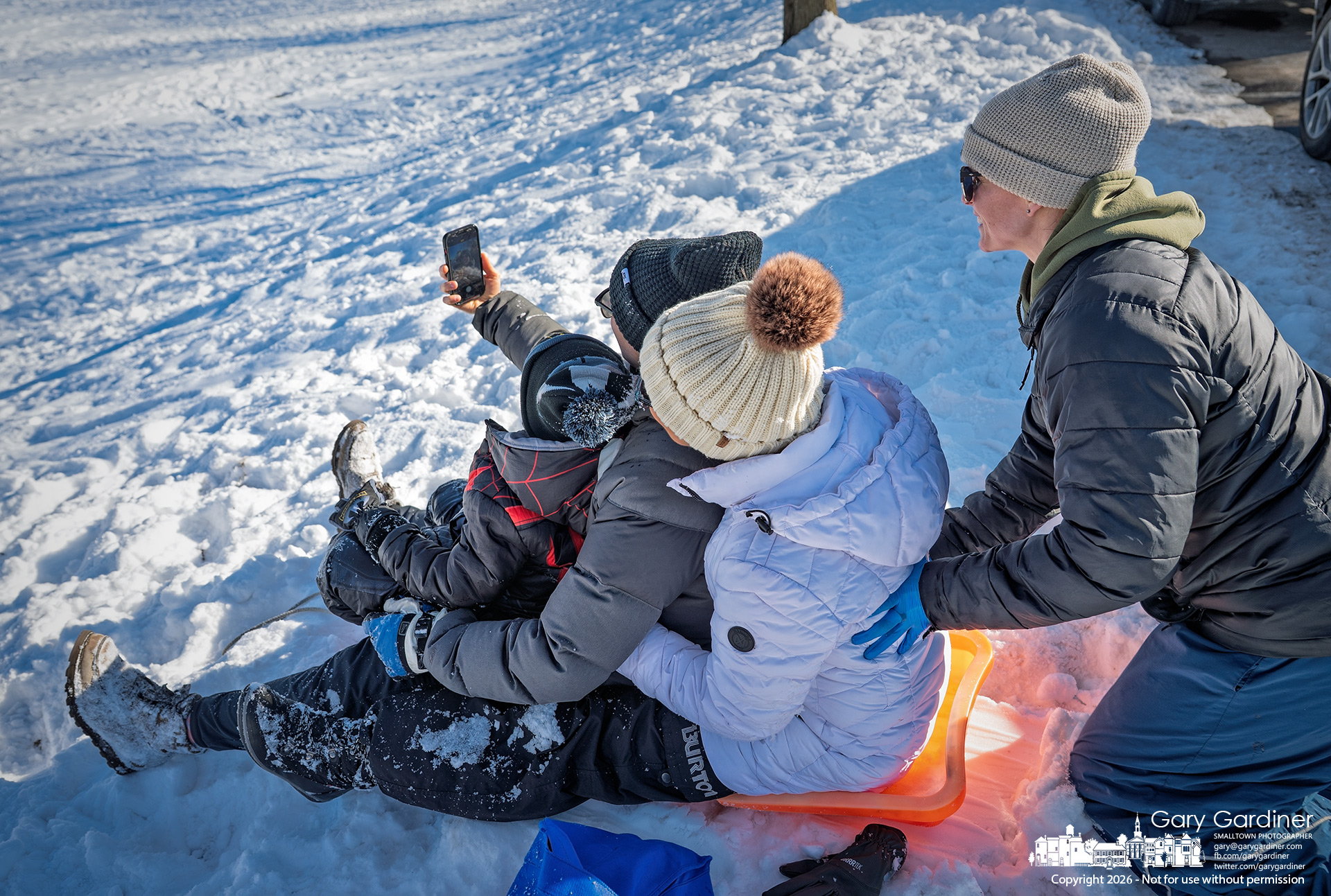 A mother holds up her phone to capture a selfie with her kids while sledding on the smaller hills at Alum Creek Park North, bundled up for a cold, snowy day. (My Final Photo for February 8, 2026)