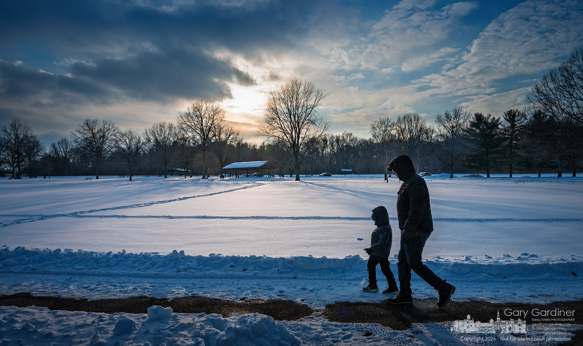 A man and a child walk along a path near Schrock Lake at Sharon Woods Park on a winter afternoon, as fresh snow covers the ground and the low sun breaks through clouds in the background. (My Final Photo for February 3, 2026)