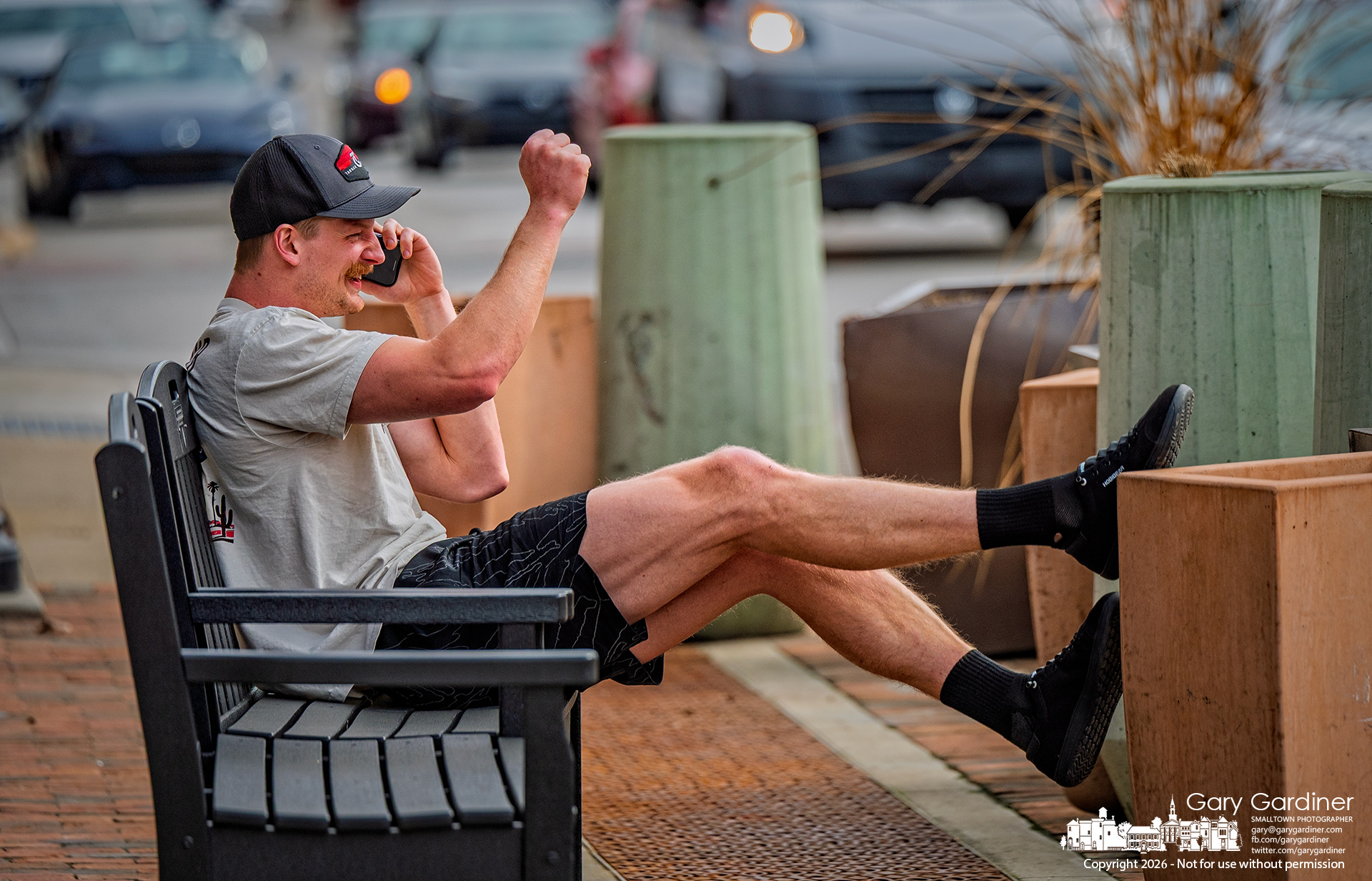 A man celebrates during a phone call in Uptown while relaxing on a bench with his feet propped on a sidewalk planter. (My Final Photo for March 25, 2026)
