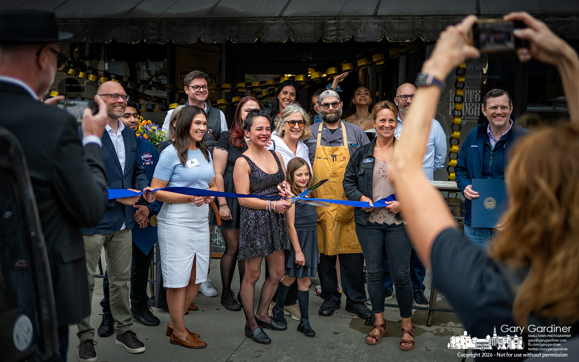 Megan Ada, at center with scissors, poses with family, friends, and others before cutting a ribbon celebrating Asterisk Supper Club's tenth anniversary Saturday afternoon. (My Final Photo for April 18, 20206 by Gary Gardiner)