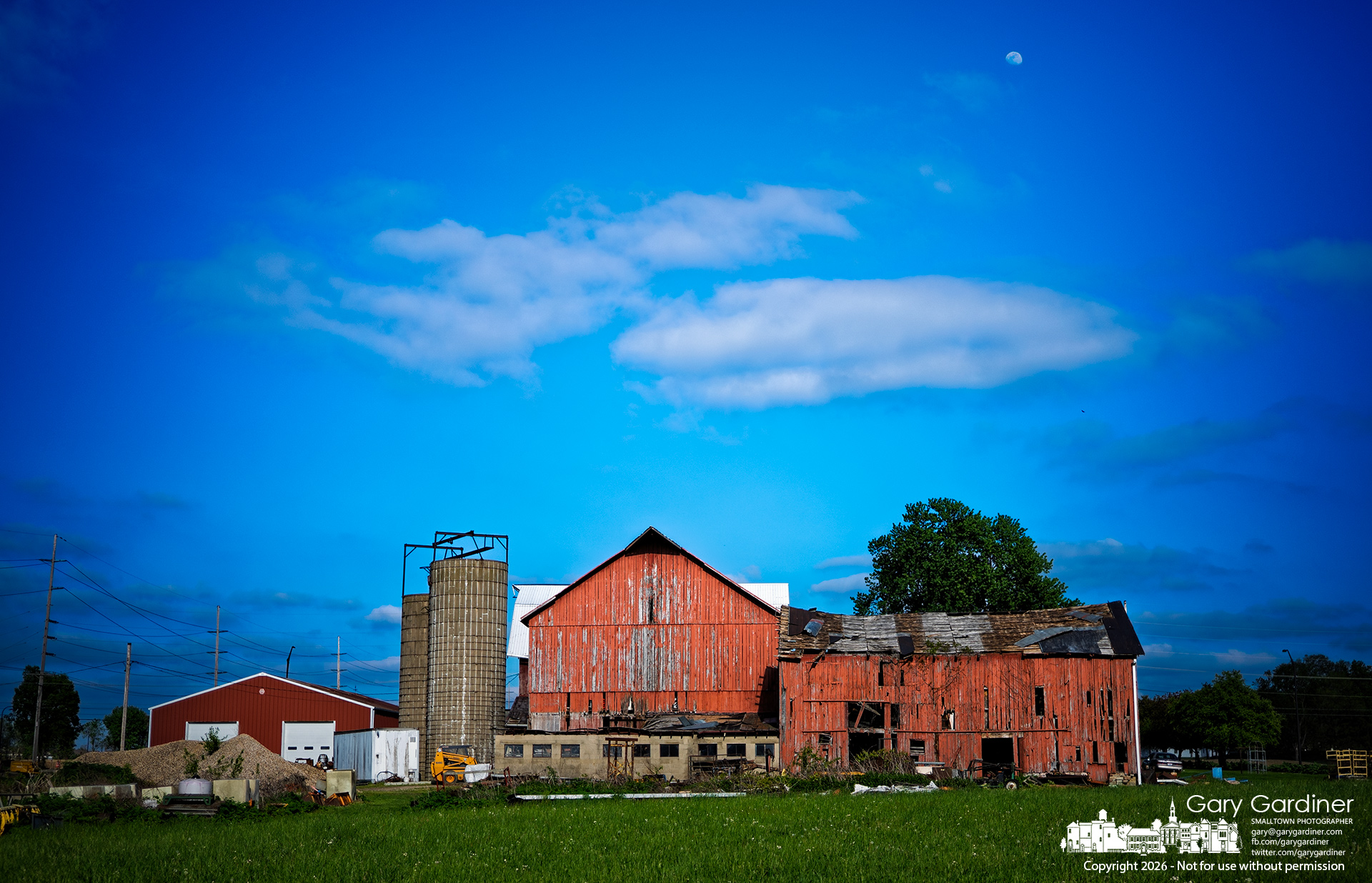 A waxing moon sits above a nearly clear sky late Sunday afternoon as the evening sun warms the barns at the Yarnell Farm on Africa Road. (My Final Photo for April 26, 2026, by Gary Gardiner)