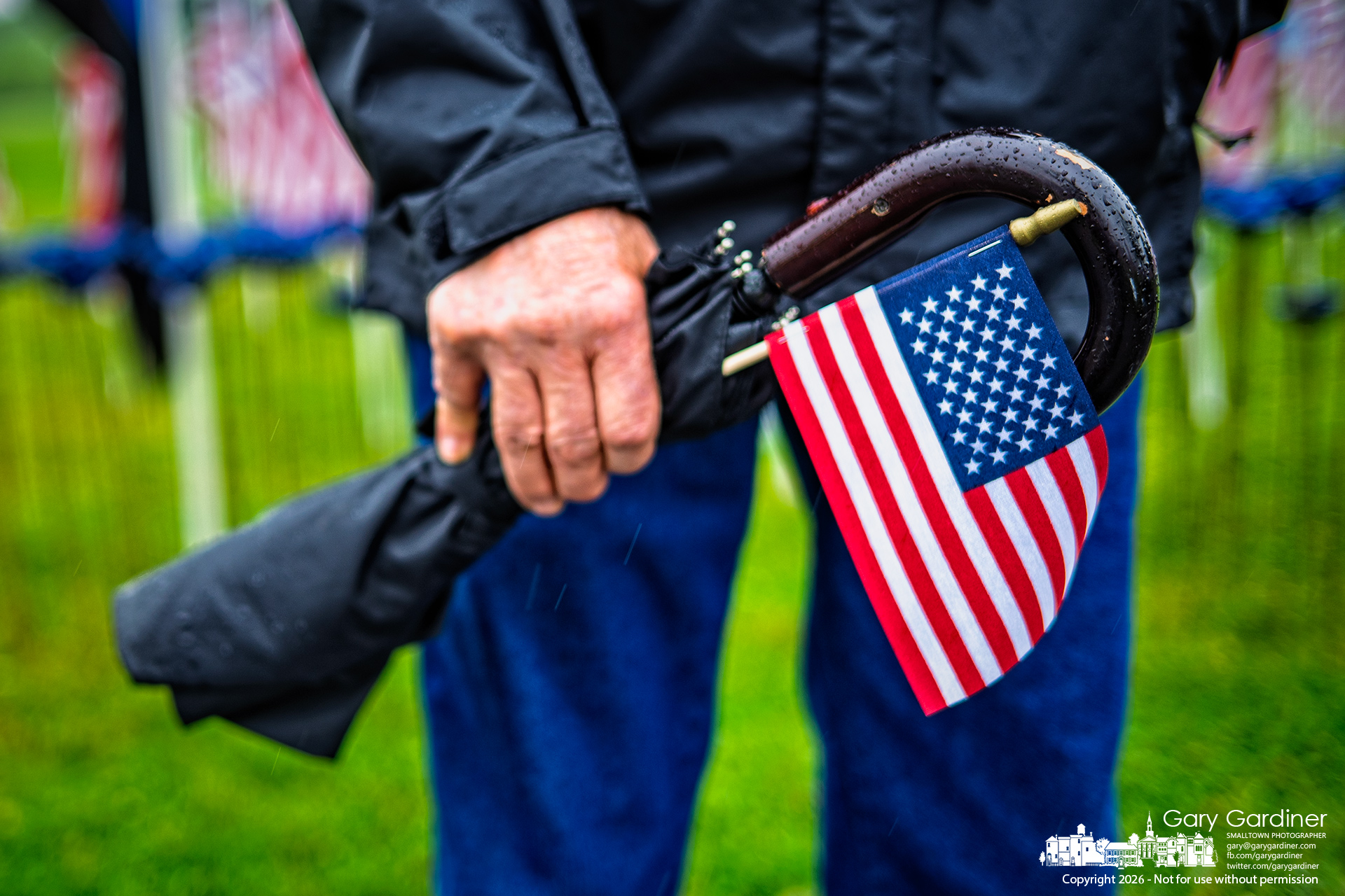 A visitor to Friday night’s opening of the Field of Heroes carries a flag and umbrella, each one designed for protection—one from the rain, the other as a symbol of the freedoms preserved by those honored in the display. (My Final Photo for May 22, 2026. Photo by Gary Gardiner)