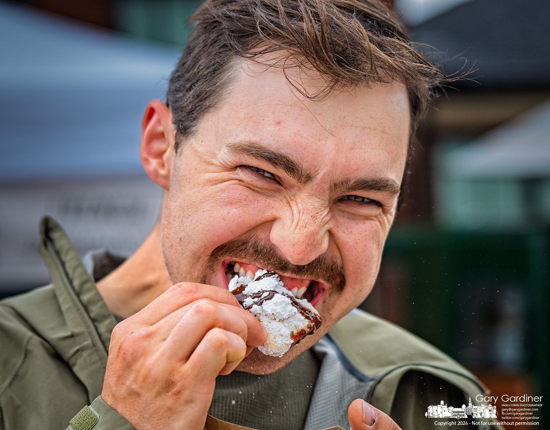 A Saturday Market shopper delicately bit into a powdered sugar-coated beignet served with coffee from a vendor on the second weekend of the summer-long market. (My Final Photo for May 23, 2026. Photo by Gary Gardiner)