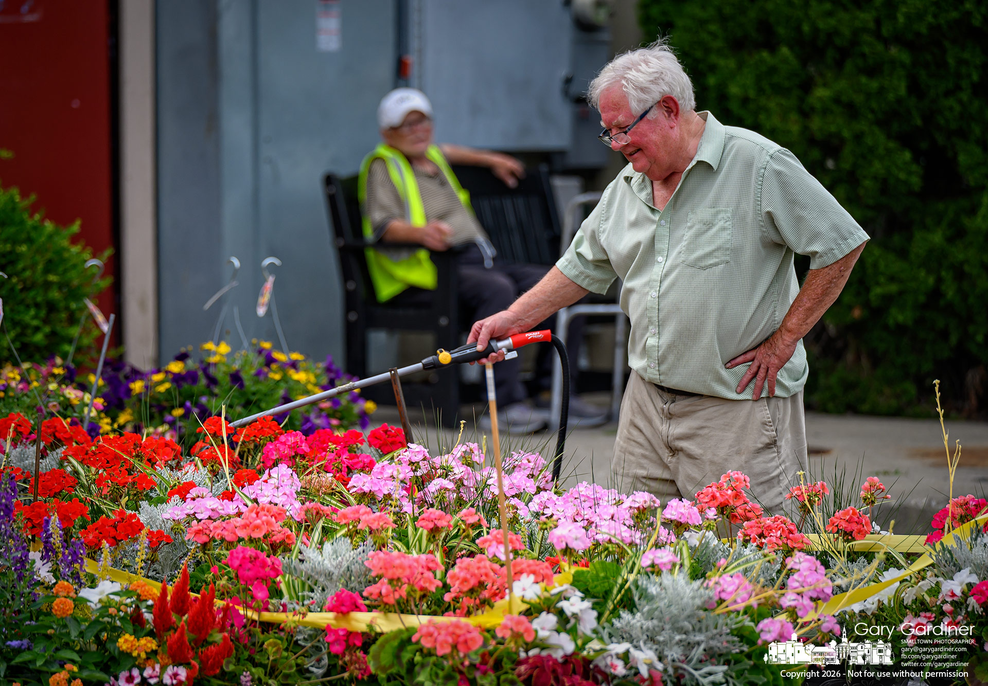 Dave Talbotts waters the flowers behind his florist shop as Uptown Dave Deringer rests in the shade by Old Bag of Nails, with florist Dave offering to cool off Uptown Dave with a sprinkler shower. (My Final Photo for June 10, 2026. Photo by Gary Gardiner)