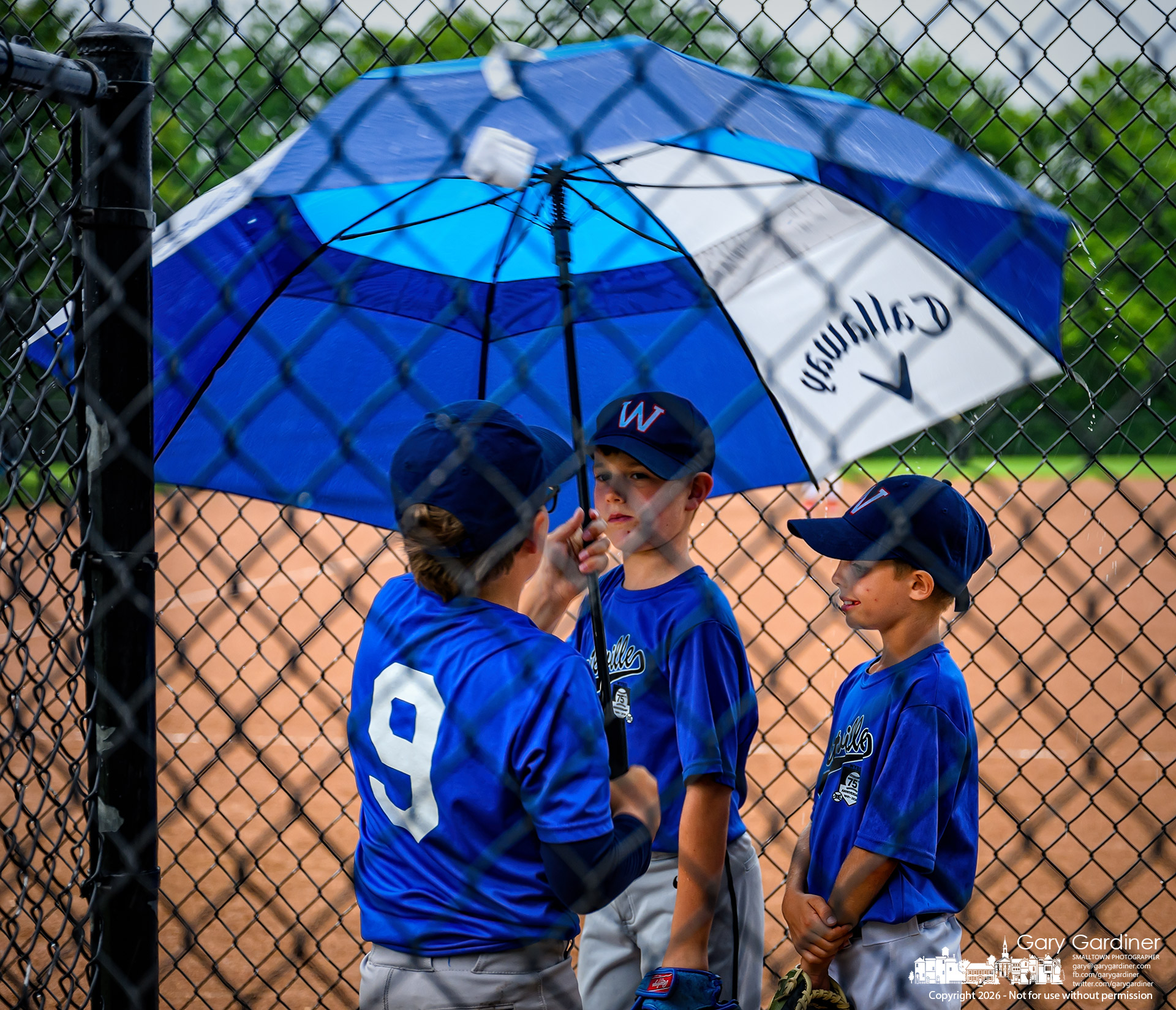 Three youth baseball players gather under a golf umbrella minutes before their game was called due to rain at Huber Village Park. (My Final Photofor June 17, 2026)