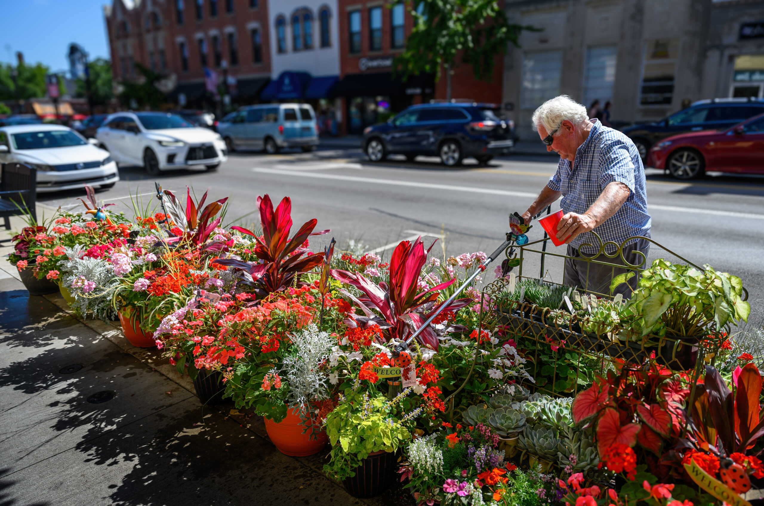 Florist Dave Talbott, with a glass of cold refreshment, waters the flowers in front of his store just before closing for the day, when the temperature reached the high 90s. (My Final Photo for July 1, 2026. Photo by Gary Gardiner)
