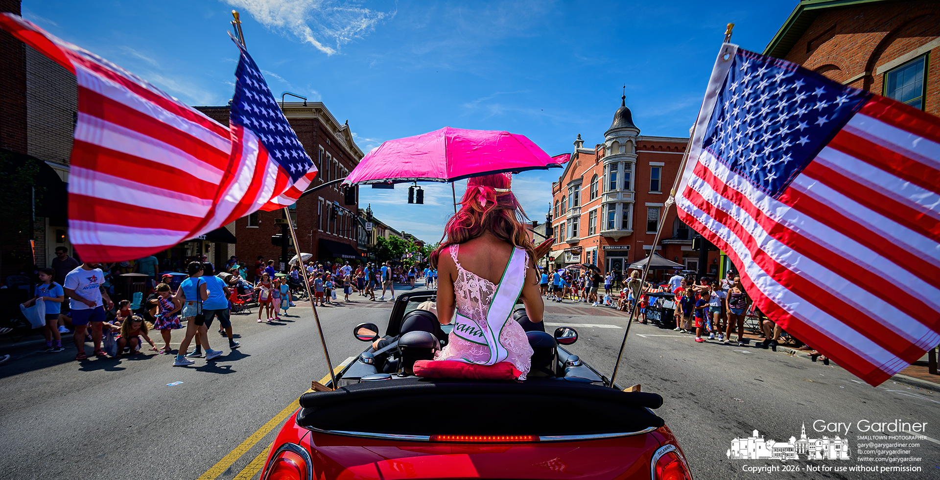 A pink umbrella shades a rider flanked by flags as they approach Uptown Westerville during the 4th of July parade. (My Final Photo for July 4, 2026. Photo by Gary Gardiner)