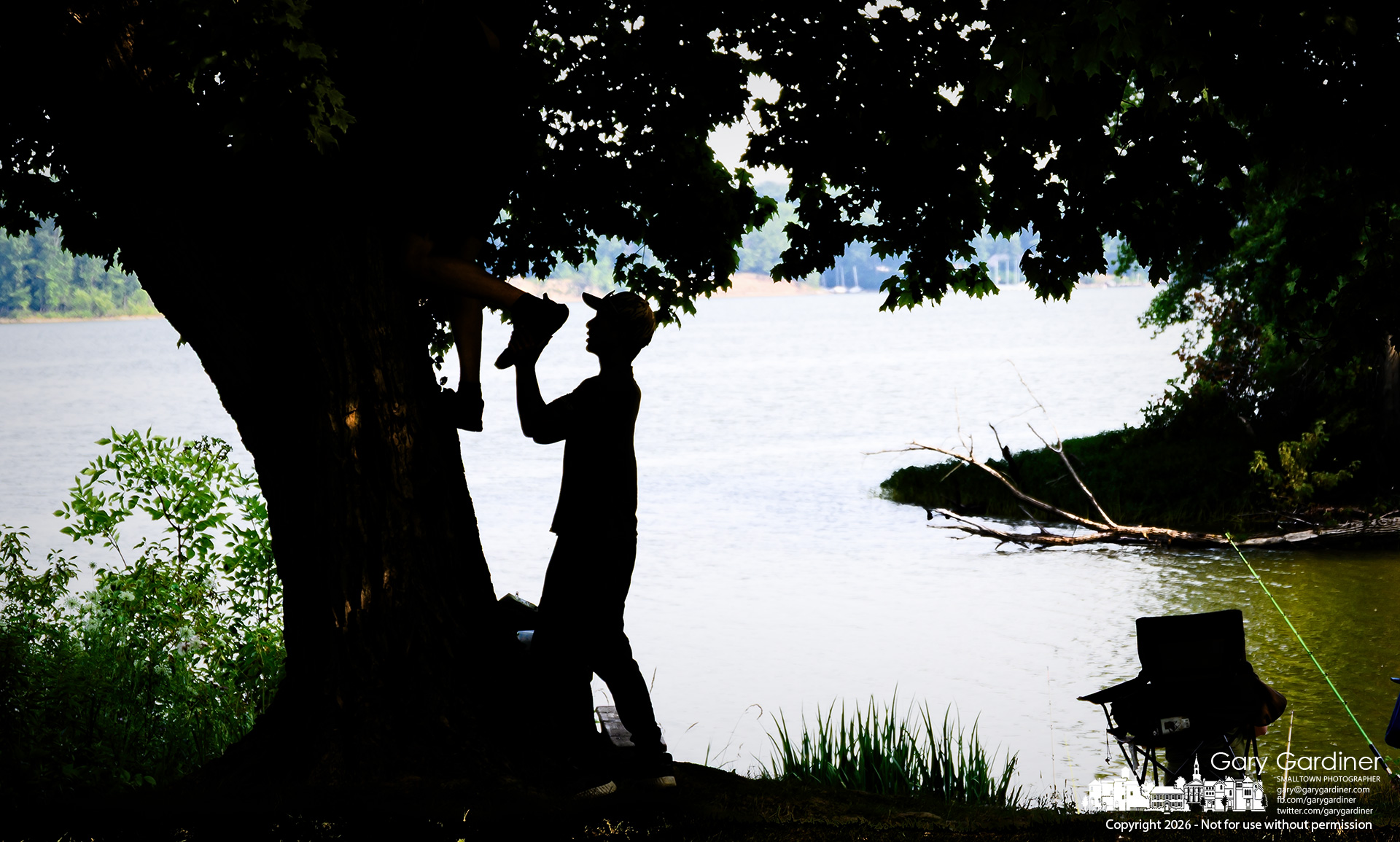 A young man helps his friend climb the mature maple tree along the shoreline of Hoover Reservoir at Red Bank Park. (My Final Photo for July 16, 2026. Photo by Gary Gardiner)