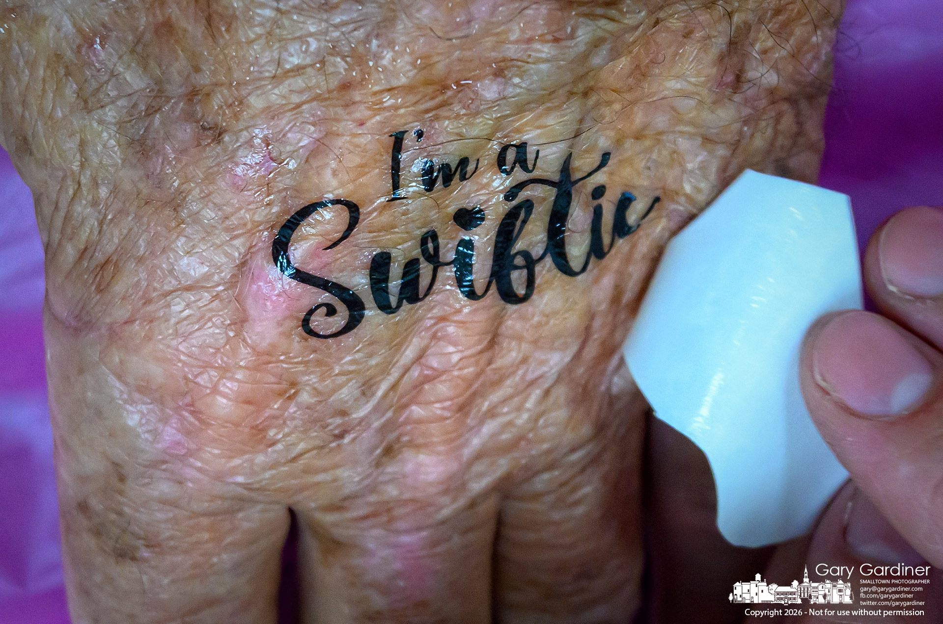 An older man, whose hand shows signs of age, gets an "I'm a Swiftie" temporary tattoo at the Taylor Swift Party at the library. (My Final Photo for July 14, 2026. Photo by Gary Gardiner)
