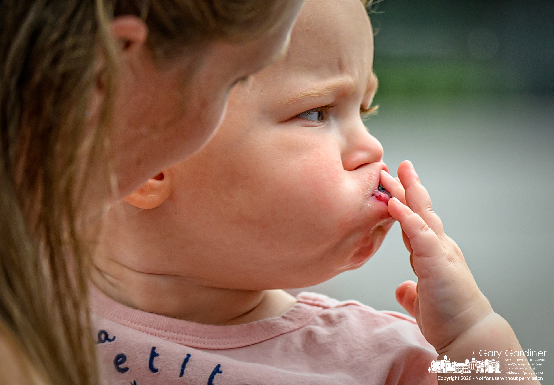 A toddler stuffs a large blackberry into her mouth before the container of berries was covered in protective netting and placed in a bag for mom to carry, ending her sweet snack at the Saturday Farmers Market. (My Final Photo for August 15, 2026. Photo by Gary Gardiner)