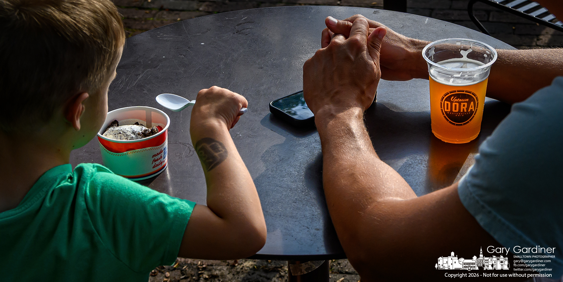 Father and son listen to musicians at Rotary Park and enjoy their choice of refreshments on a warm Friday evening in Uptown. (My Final Photo for August 14, 2026. Photo by Gary Gardiner)