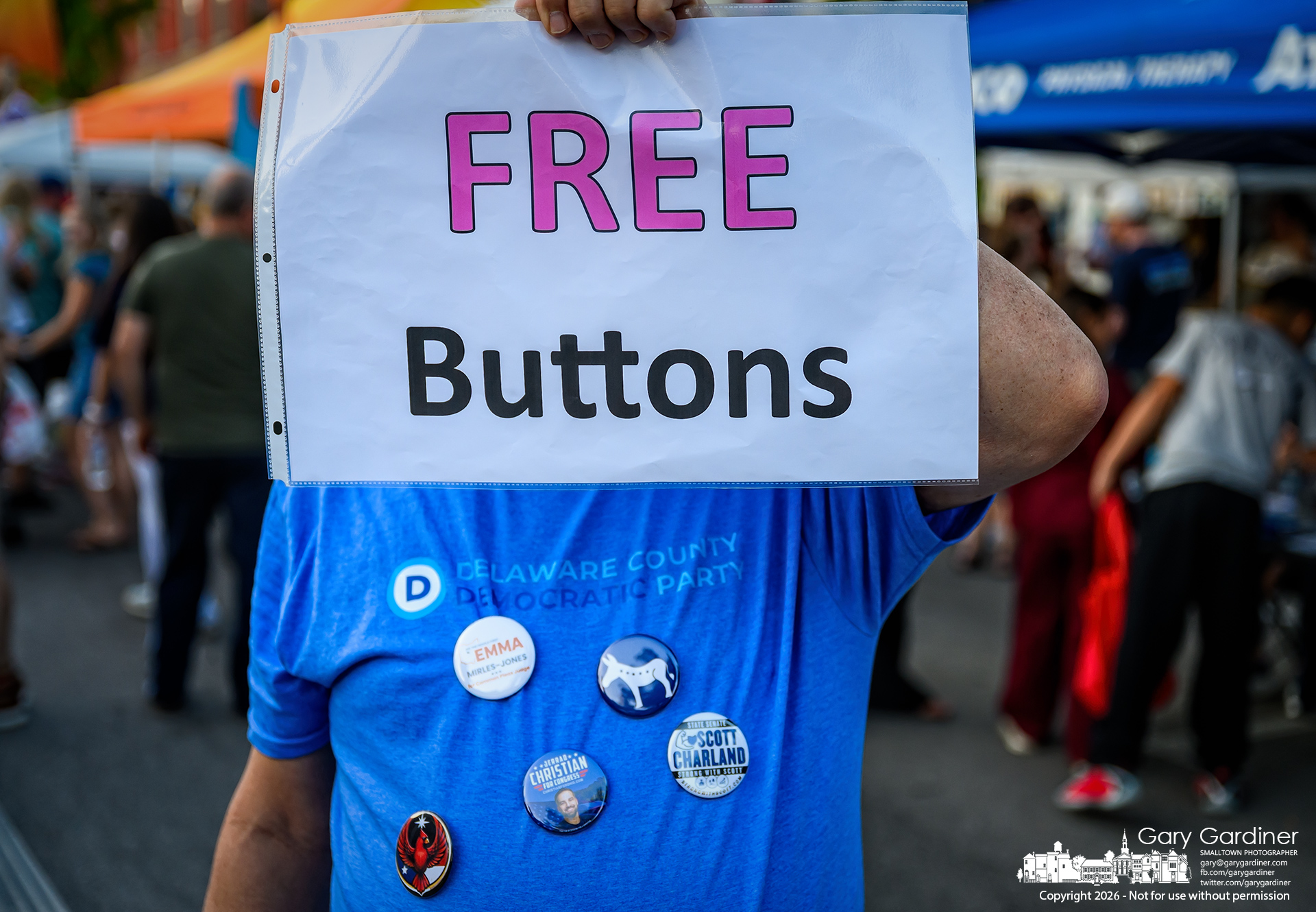 A canvasser holds up a sign offering free campaign buttons at a booth at Uptown Westerville's Fourth Friday. (My Final Photo for August 28, 2026. Photo by Gary Gardiner)