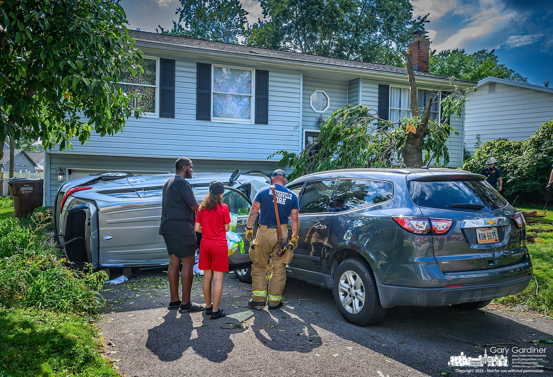 A Westerville firefighter talks with a father and daughter after their car, parked in their driveway on Paris Blvd. in Blendon Township, was struck by a car that failed to navigate a turn, left the roadway, struck a tree in the front yard, flipped onto its side, and landed between their car and the garage door. (My Final Photo for September 9, 2026. Photo by Gary Gardinner)