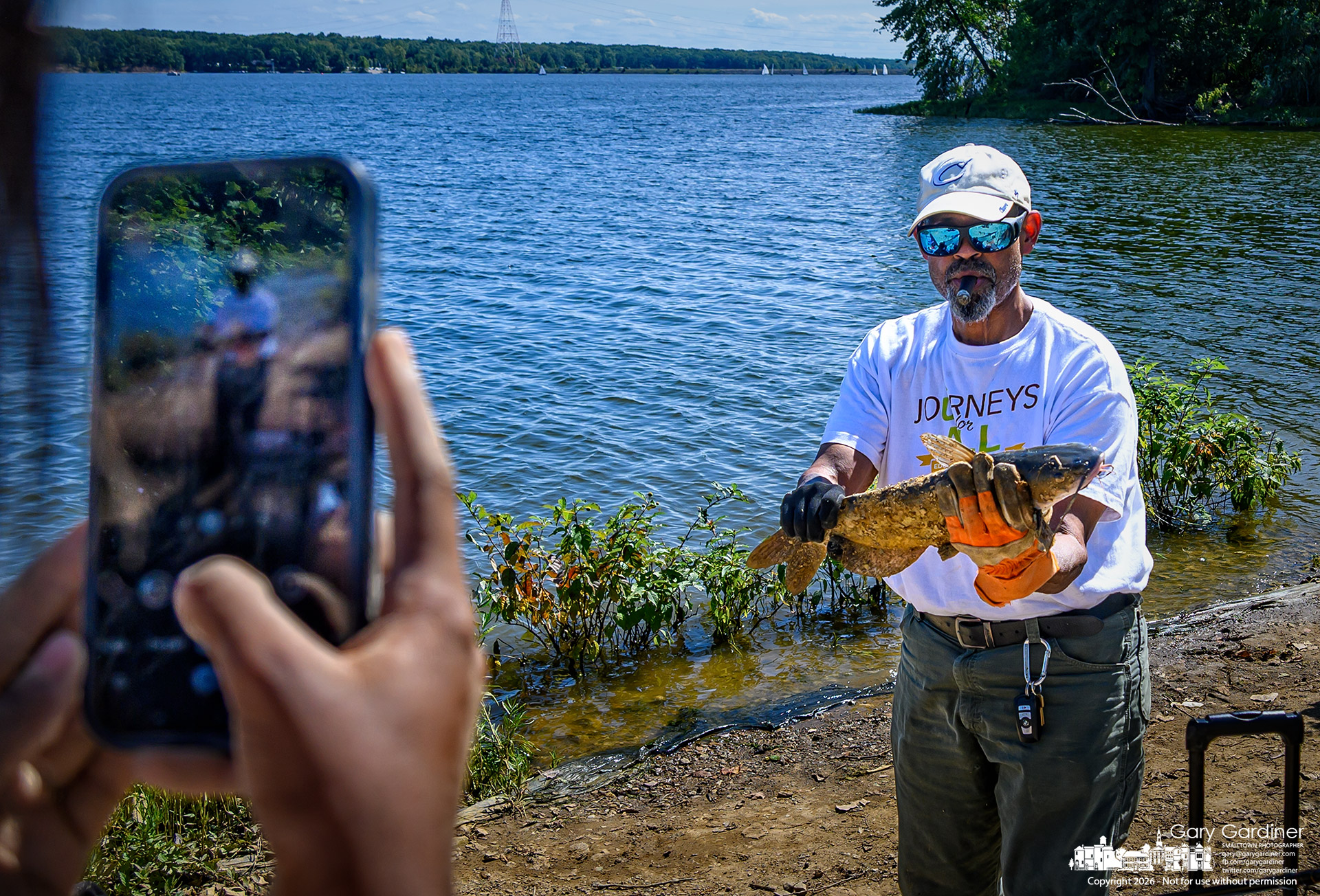 A cigar-smoking fisherman poses for a photo with the second catfish he pulled from Hoover Reservoir at Red Bank Park on Labor Day. (my Final Photo for September 7, 2026. Photo by Gary Gardiner)