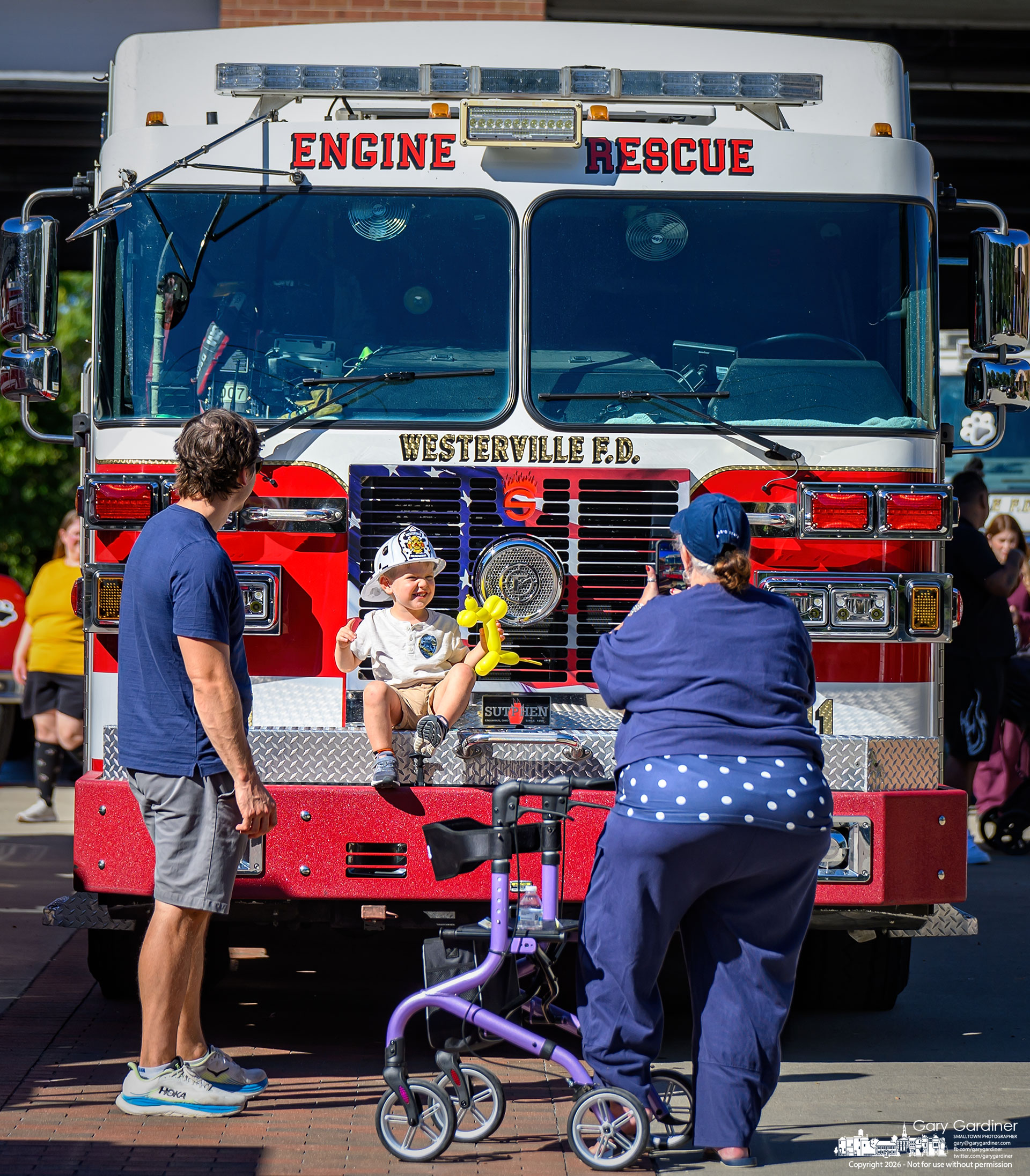 A cheerful child rests on the bumper of a fire truck while his parents photograph him at the Fire Prevention Open House at the main fire station on West Main Street. (My Final Photo for October 4, 2026. Photo by Gary Gardiner)