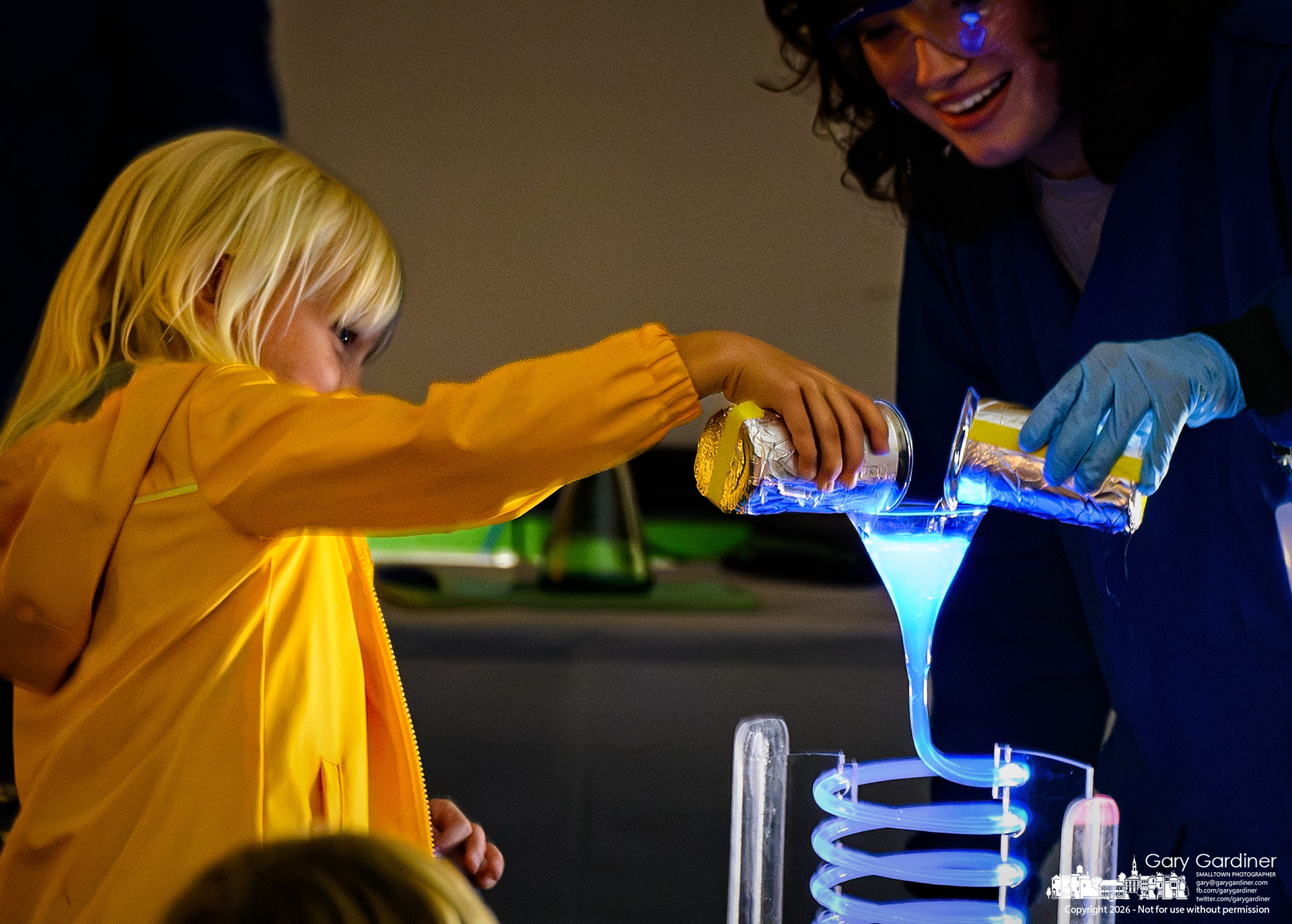 A young scientist assists an older counterpart with an experiment that made two chemicals fluoresce during the OSU Magic of Science demonstration at the library.(My Final Photo for October 16, 2026. Photo by Gary Gardiner)